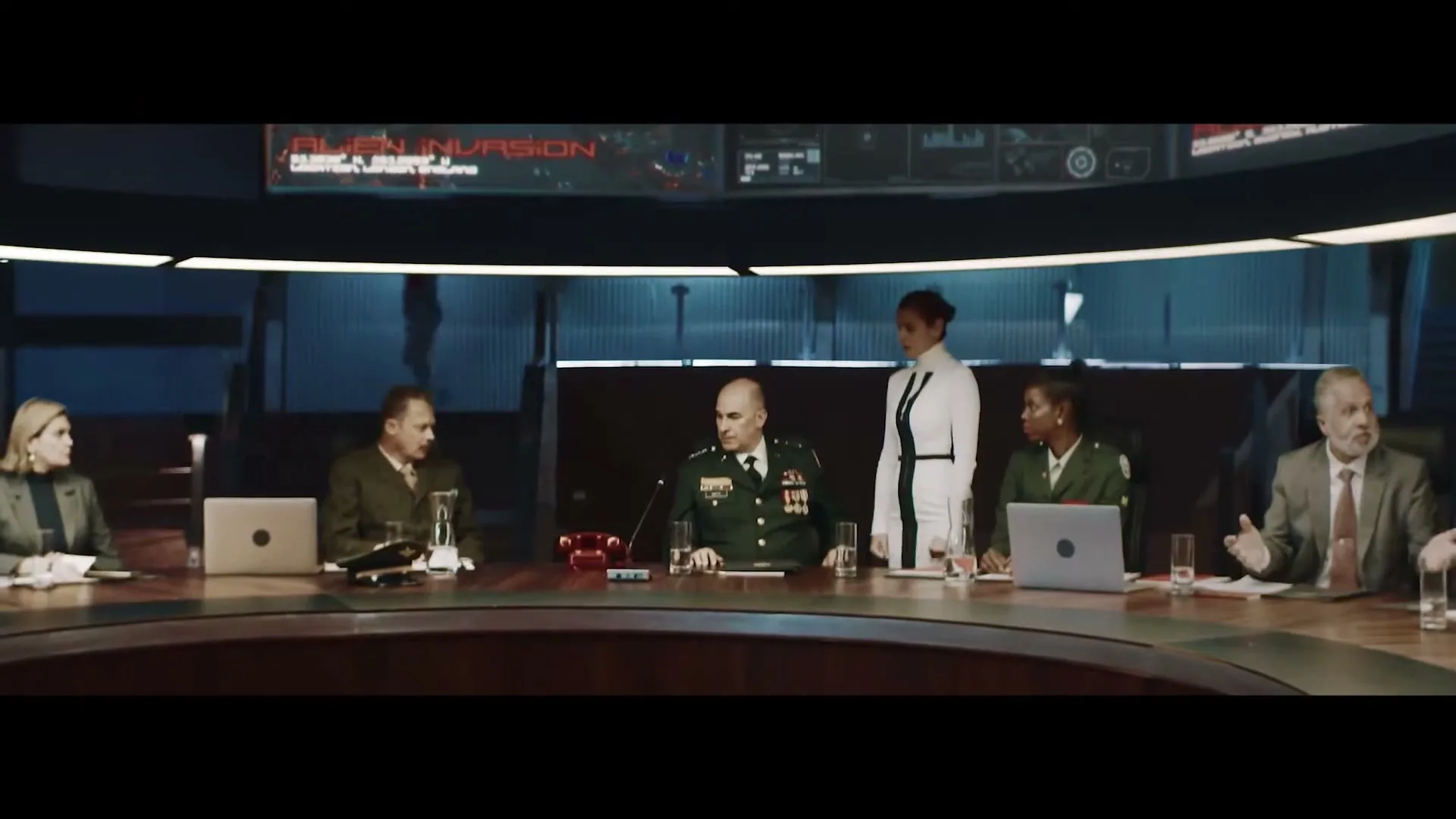 A diverse group of military and civilian leaders are seated around a large, curved conference table in a dimly lit, high-tech command center, discussing an alien invasion.