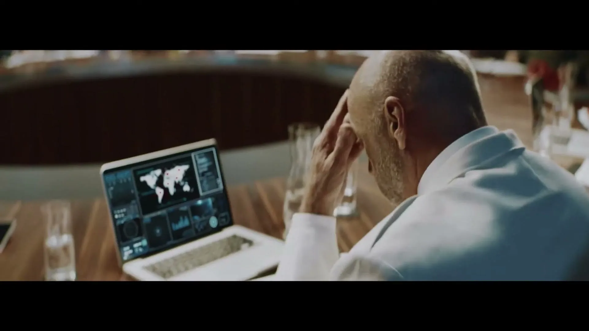A bald, older man in a white shirt sits at a large conference table, looking stressed while viewing a laptop displaying a world map with red dots.