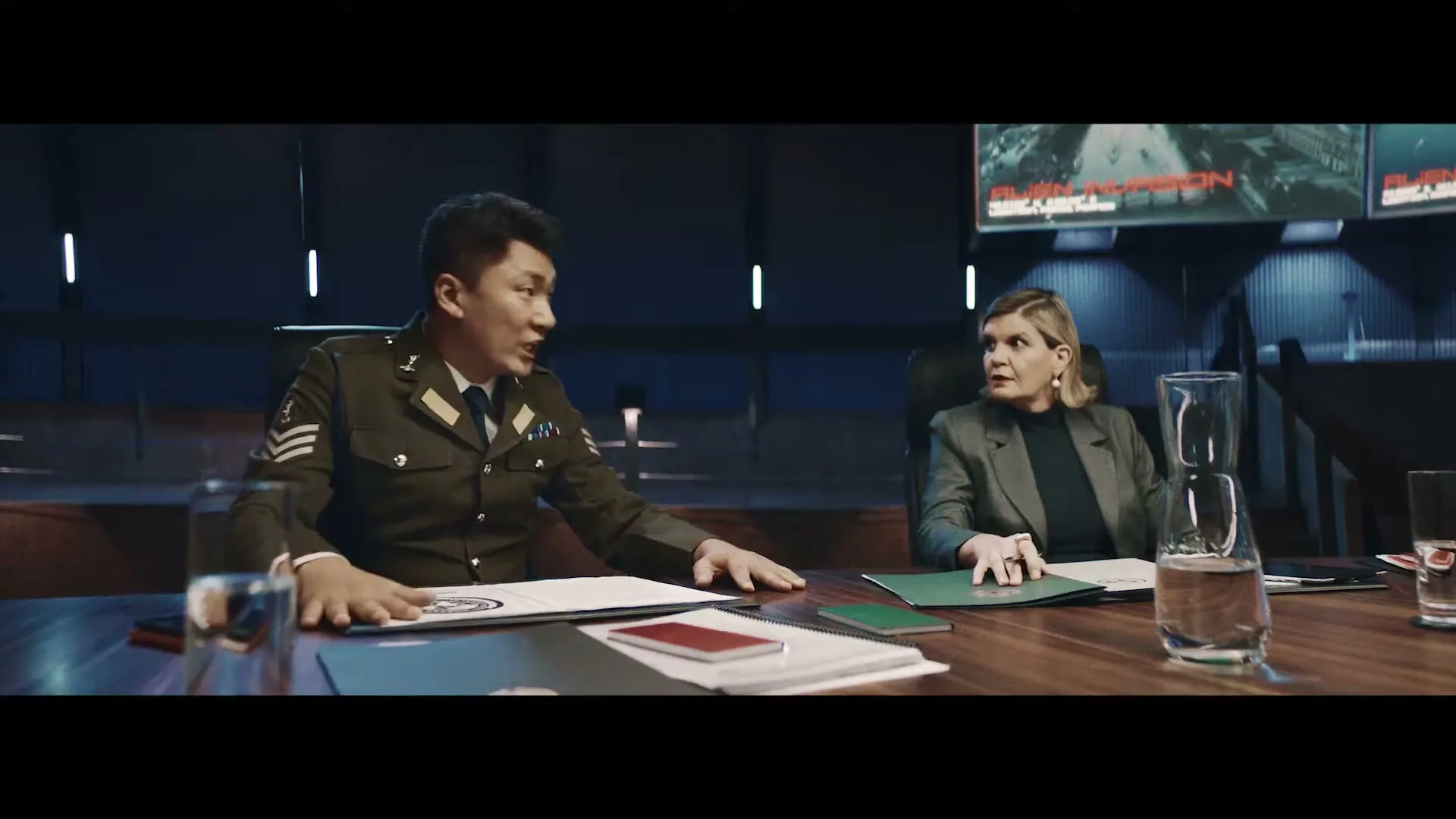 A male general in uniform and a female executive in a blazer sit at a large conference table, discussing documents in a dimly lit, modern meeting room.
