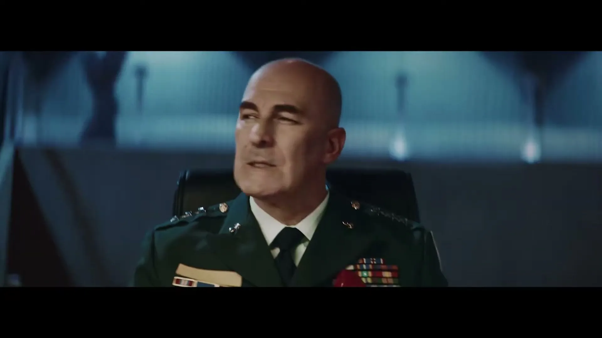 A bald male general in a green uniform with medals sits at a table, looking around a dark, futuristic command center.