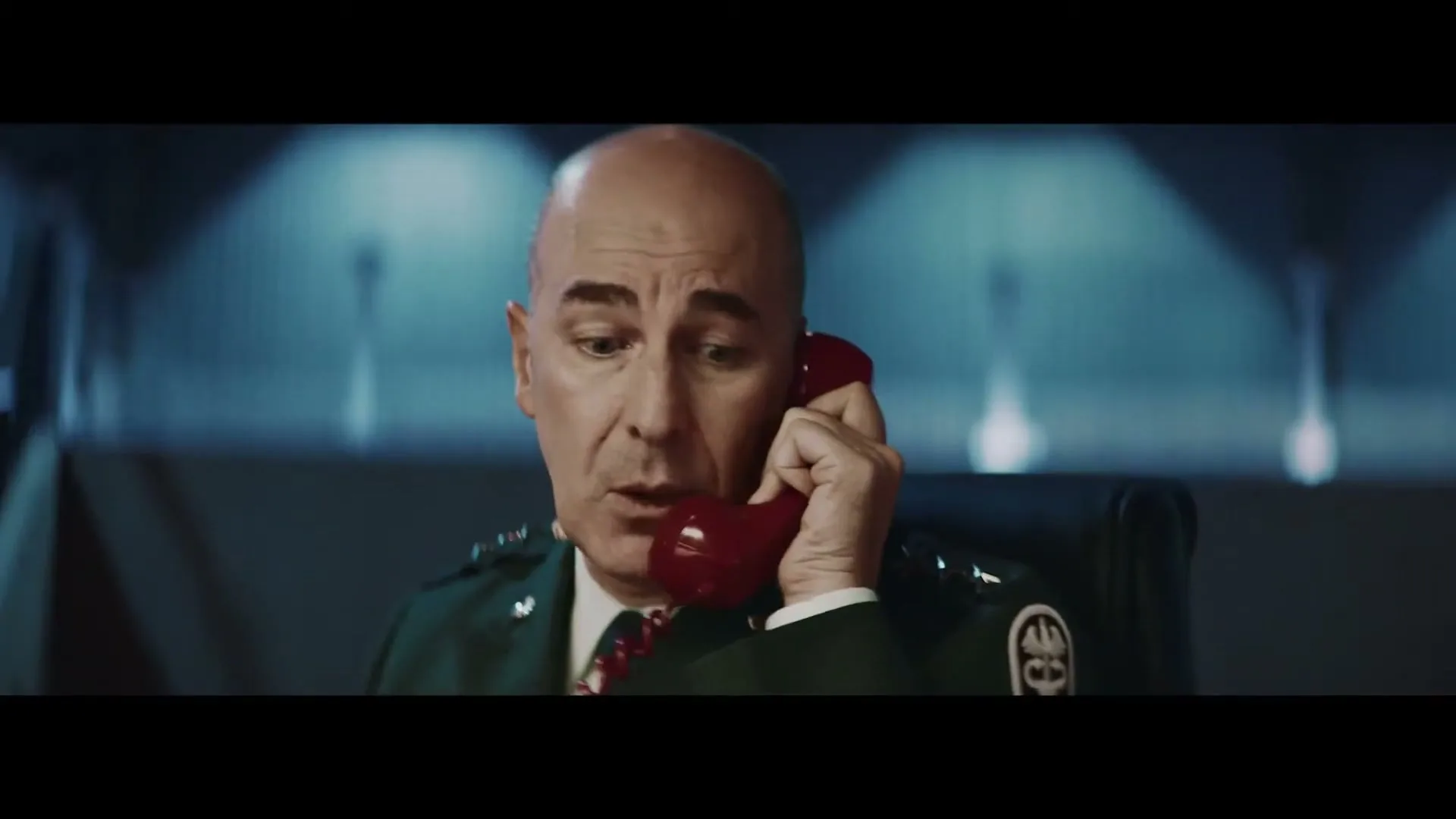 A bald man in a green military uniform holds a red phone to his ear, looking concerned in a dimly lit control room.