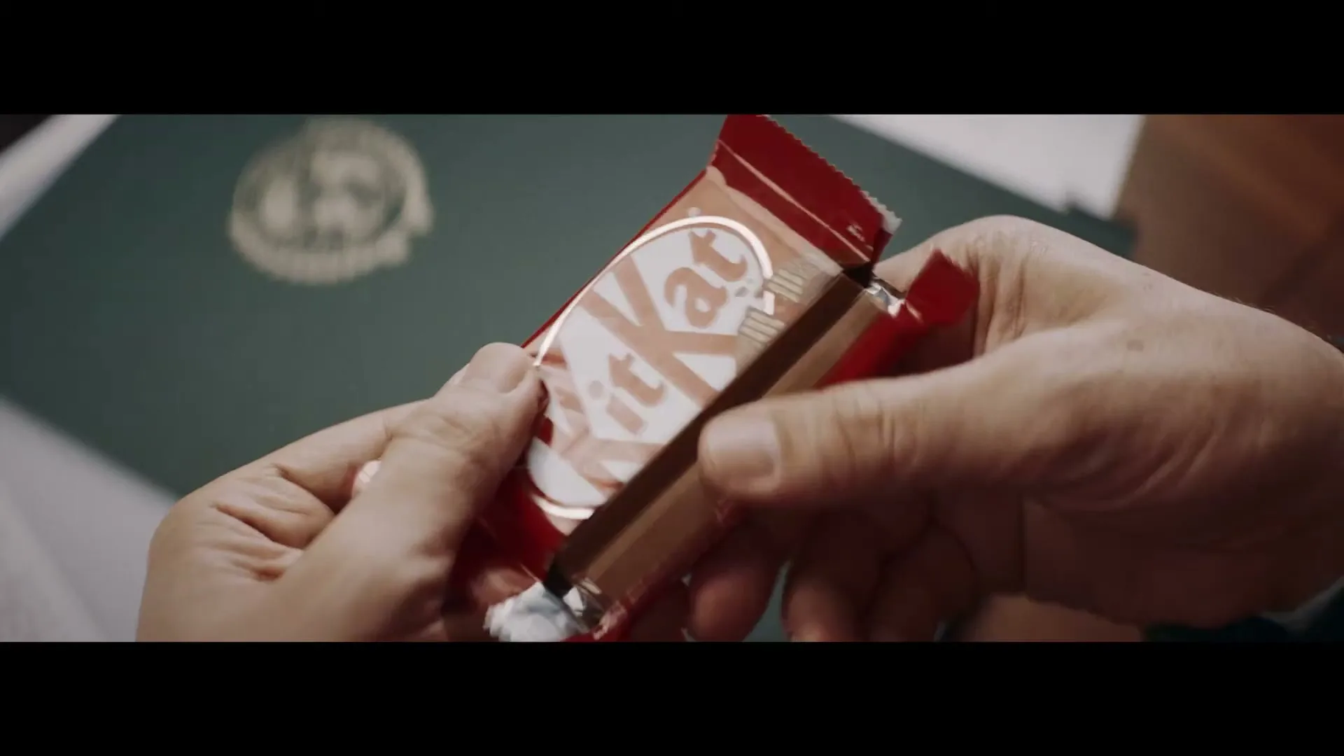 Two hands, one with a dark suit sleeve, hold a red Kit Kat bar, unwrapping it to reveal the chocolate inside, against a blurred green background.