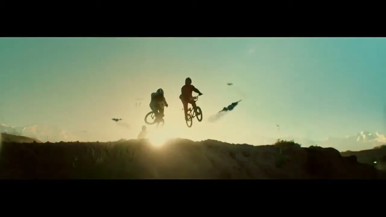 Two silhouetted BMX riders jump over a dirt mound against a bright sky, with lens flare and flying objects.