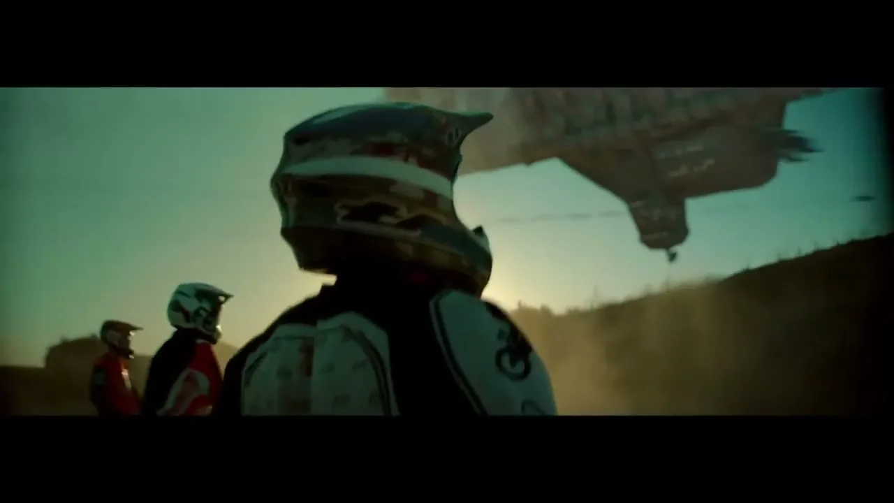 Three motocross riders in full gear stand on a dusty terrain, looking up at a large, dark spaceship hovering in the sky during sunset.