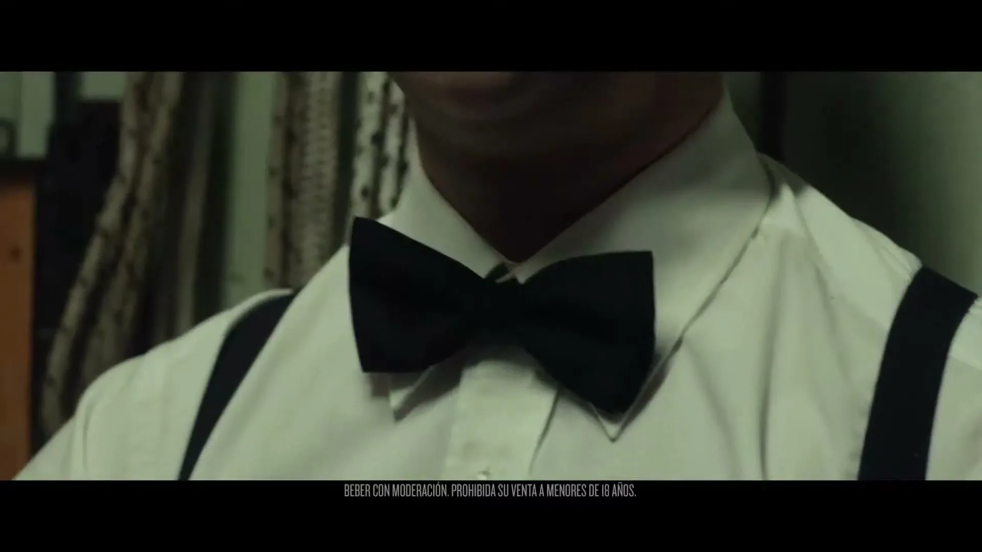 A man in his 30s, wearing a white shirt, black bow tie, and suspenders, is seen from the chest up, adjusting his bow tie in an interior setting.