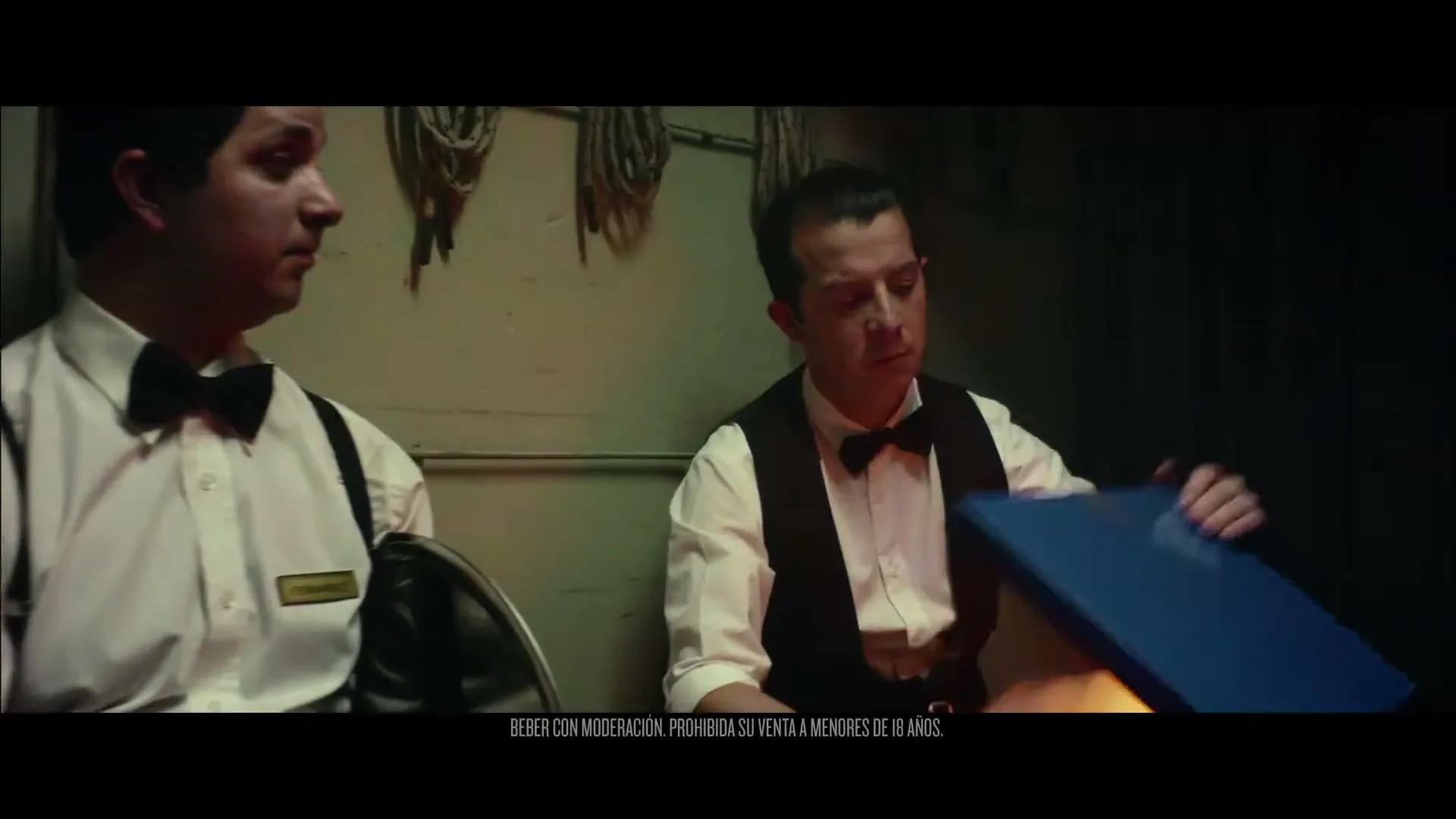 Two male waiters in bow ties and vests sit in a dimly lit backroom, one opening a cooler to reveal a glowing orange light and a beer bottle.