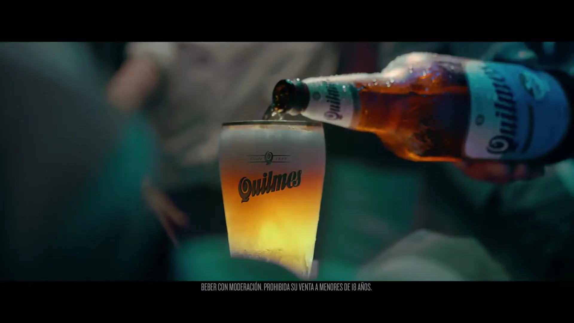 A hand pours a Quilmes beer from a bottle into a glass, creating a frothy head, in a dimly lit bar.