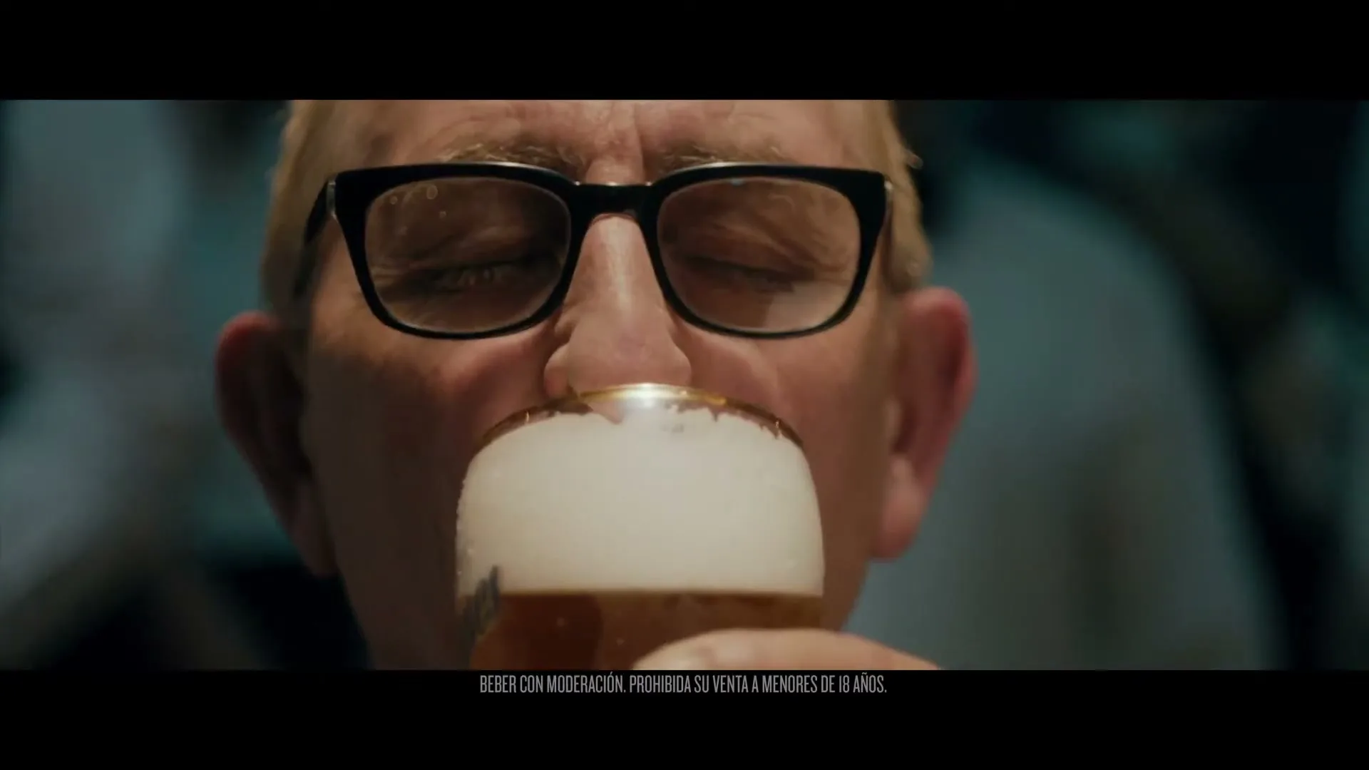 A senior man with glasses drinks a beer, eyes closed in enjoyment, in a dimly lit interior with other people blurred in the background.