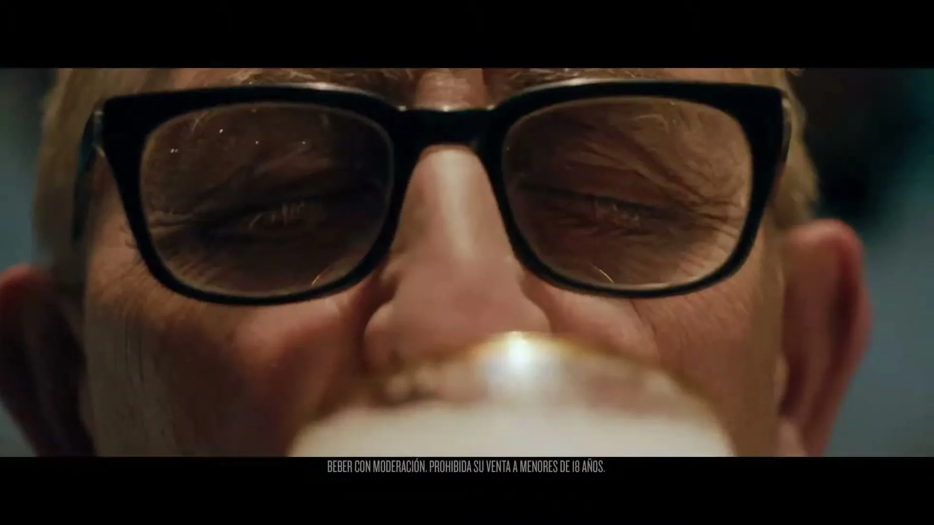 A close-up shot of an older man with black-rimmed glasses, eyes closed, as he drinks from a glass, with light reflecting off his face.
