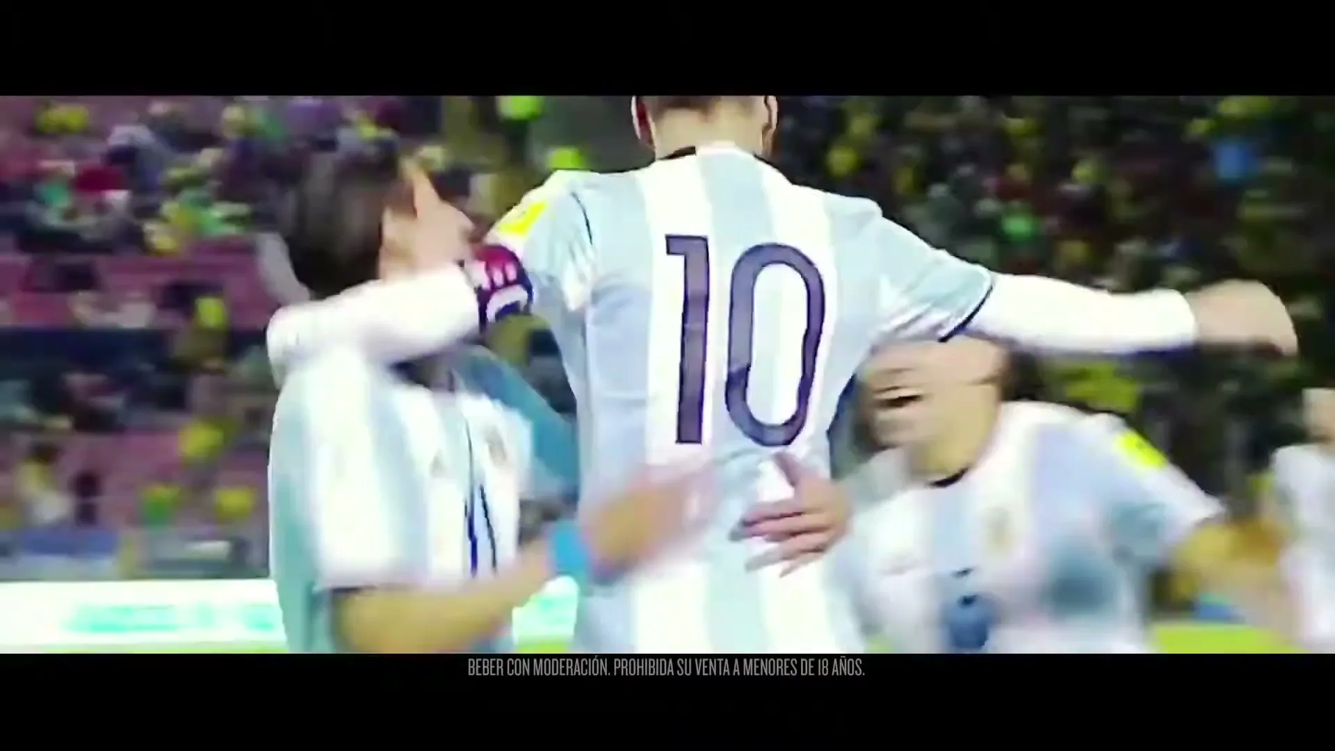 Three male soccer players in light blue and white jerseys celebrate a goal, hugging each other on a vibrant green field in a stadium.