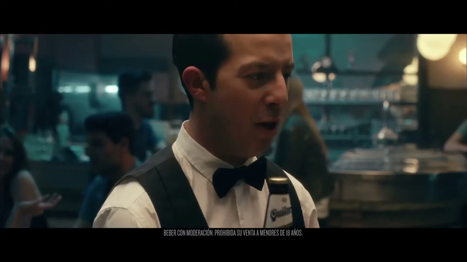 A male waiter in a white shirt and black vest holds a beer bottle and speaks to someone off-camera in a dimly lit bar.