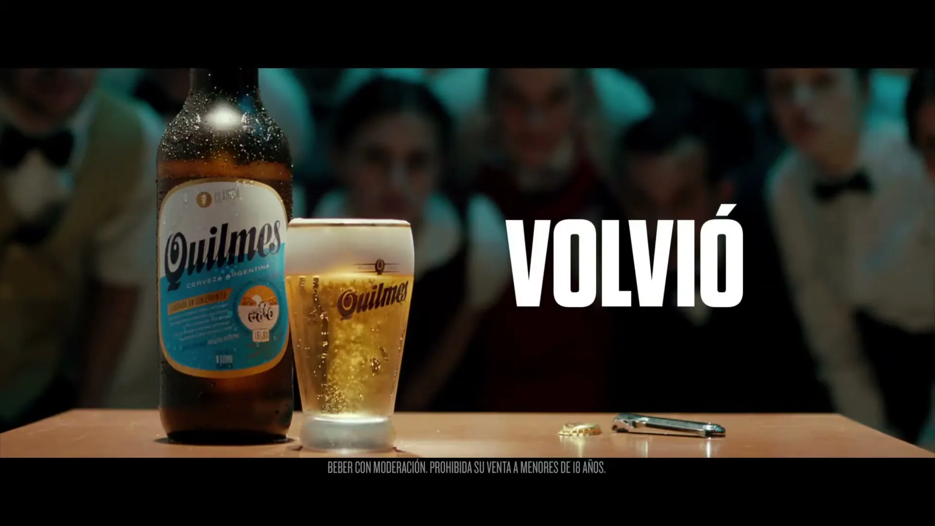 A bottle of Quilmes beer and a glass of beer sit on a wooden table, with blurred people in the background and text appearing on screen.