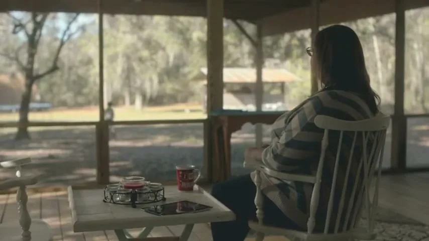 A woman with long dark hair and glasses sits in a rocking chair on a screened porch, looking out at a sunny, wooded landscape.