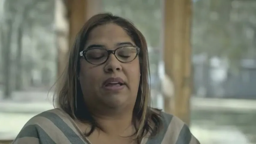 A Hispanic woman in her 40s, wearing glasses and a striped top, speaks directly to the camera in a medium shot, with a blurred outdoor background.