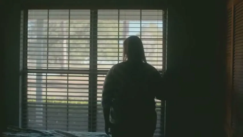 A woman stands silhouetted against a window with blinds, looking outside from a dimly lit room.