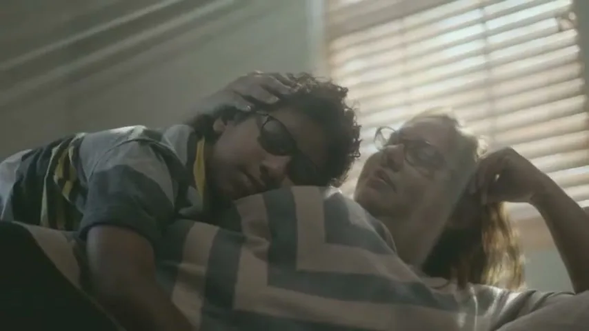 A young boy with curly hair and glasses rests his head on a woman's chest, who is also wearing glasses, as they relax on a couch indoors.