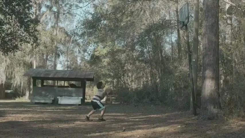 A young boy in a striped shirt and shorts plays basketball alone on a dirt court in a wooded area during the day.