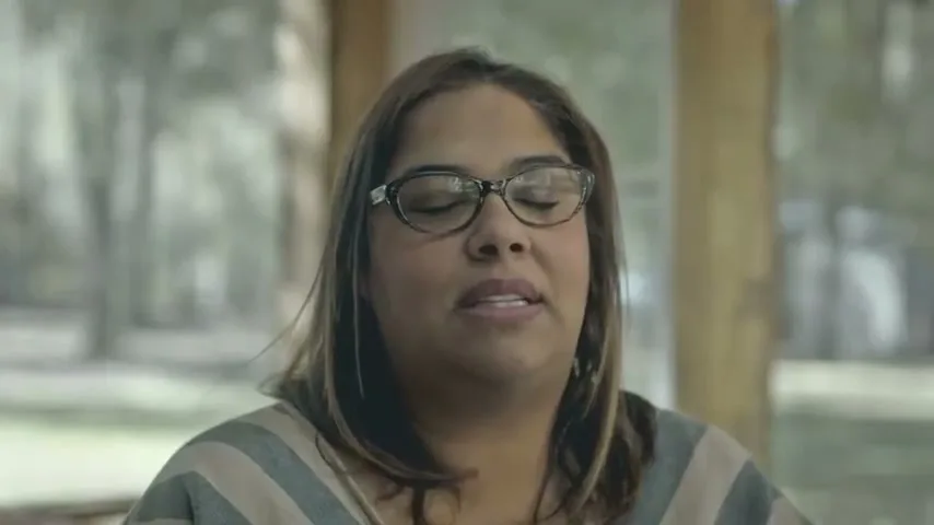 A woman with glasses and a striped top speaks to the camera, her eyes closing and opening, in an outdoor setting with trees in the background.
