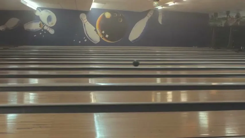 A bowling ball rolls down a lane towards the pins, with a mural of bowling pins and a ball on the wall in the background.