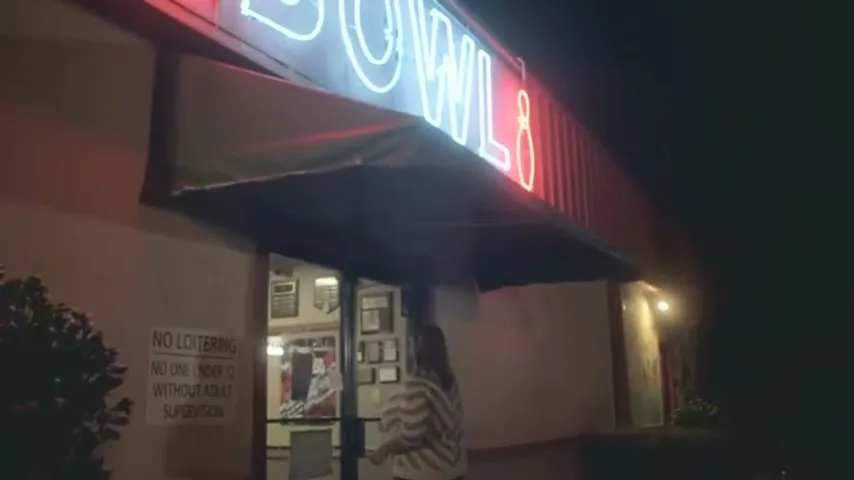 A young woman with long brown hair, wearing a striped shirt, walks towards the entrance of a bowling alley at night.