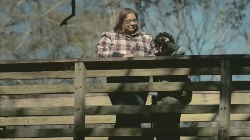 A woman in a plaid shirt and glasses stands on a wooden bridge with a child, who is looking through binoculars, in a natural outdoor setting.