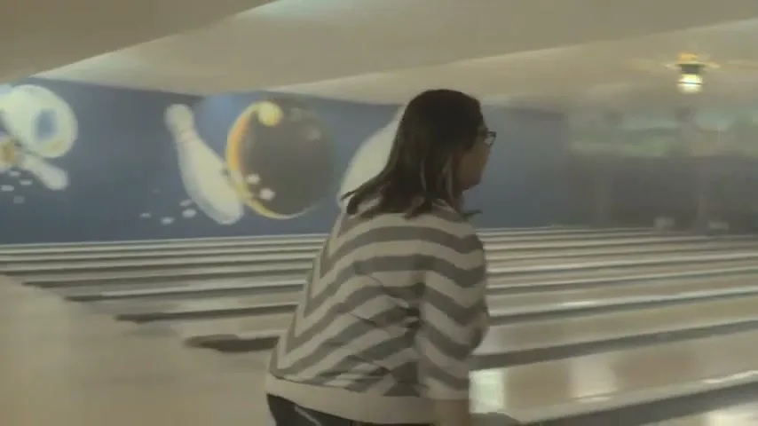 A young boy with curly hair and glasses bowls in a brightly lit bowling alley, his arm extended as he releases the ball.