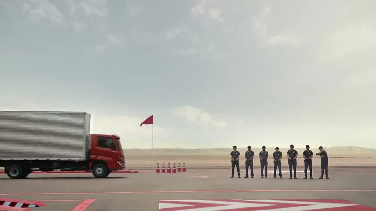 A red truck with a white cargo container drives past a line of seven men in uniform on a desert test track under a cloudy sky.