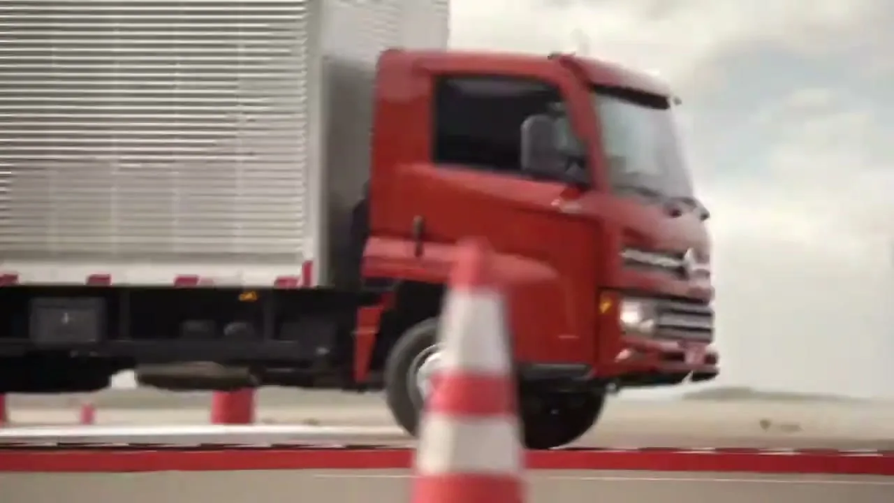 A red Volkswagen truck with a silver cargo bed drives on a test track, navigating over a red speed bump under a cloudy sky.