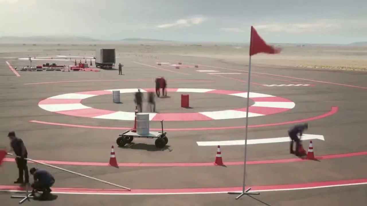 A wide shot shows a group of crew members setting up a test track with cones and barrels on an asphalt surface in a desert landscape during the day.
