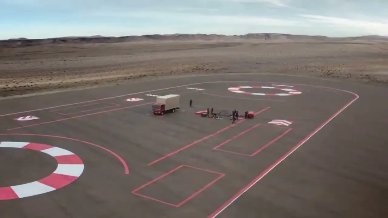 An aerial view shows a red truck and a small group of people on a test track in a desert landscape.