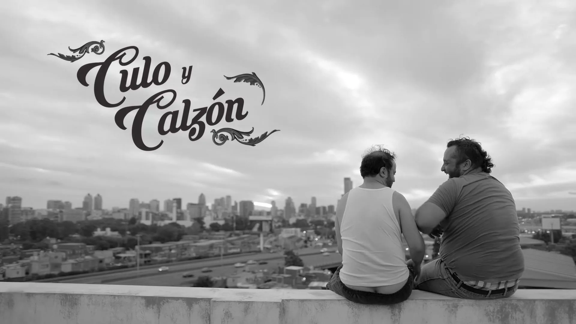 Two men sit on a rooftop ledge, looking at each other and smiling, with a city skyline in the background under a cloudy sky.