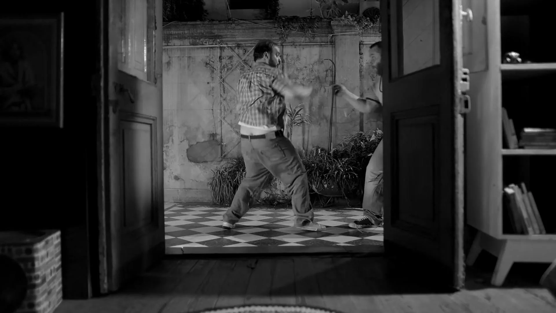 Two men, one in a plaid shirt and the other in a t-shirt, are seen through a doorway, playfully fighting in a courtyard with a checkered floor.