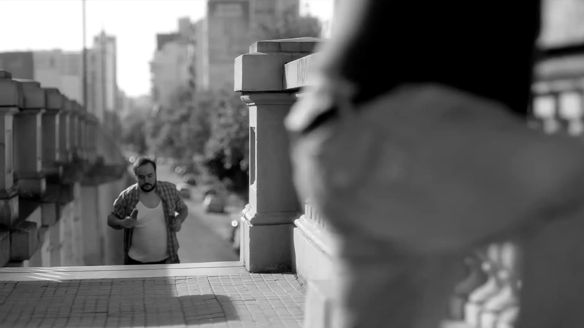A man in a plaid shirt and tank top walks up a set of stairs on a city street, looking at someone off-camera.