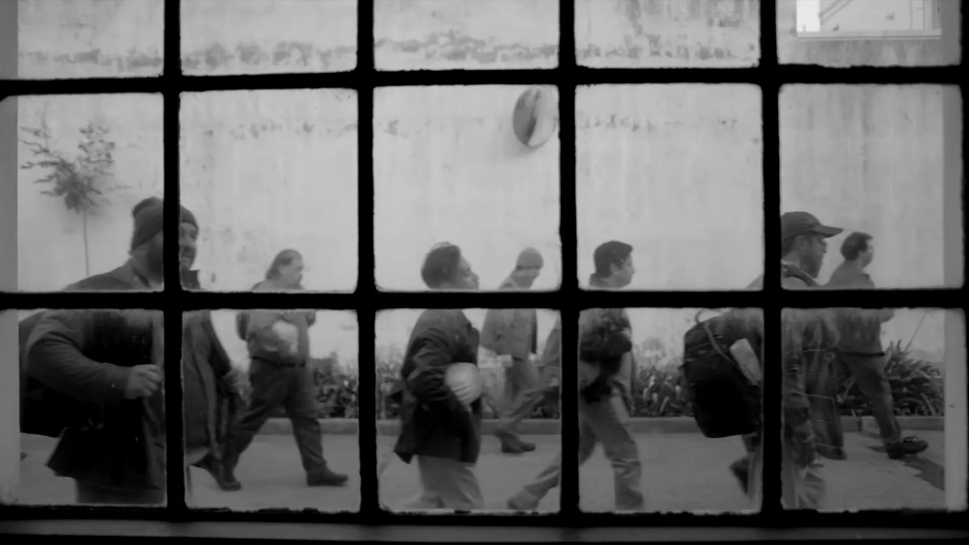A group of men walk past a window, seen through the grimy panes, in black and white, with a wall and plants in the background.