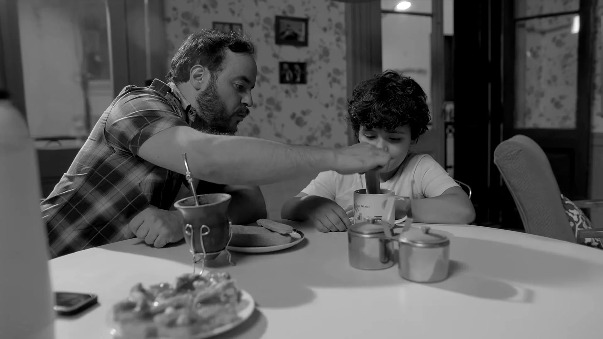 A father with a beard and a plaid shirt helps his young son stir a drink at a kitchen table, with food and a mate cup nearby.