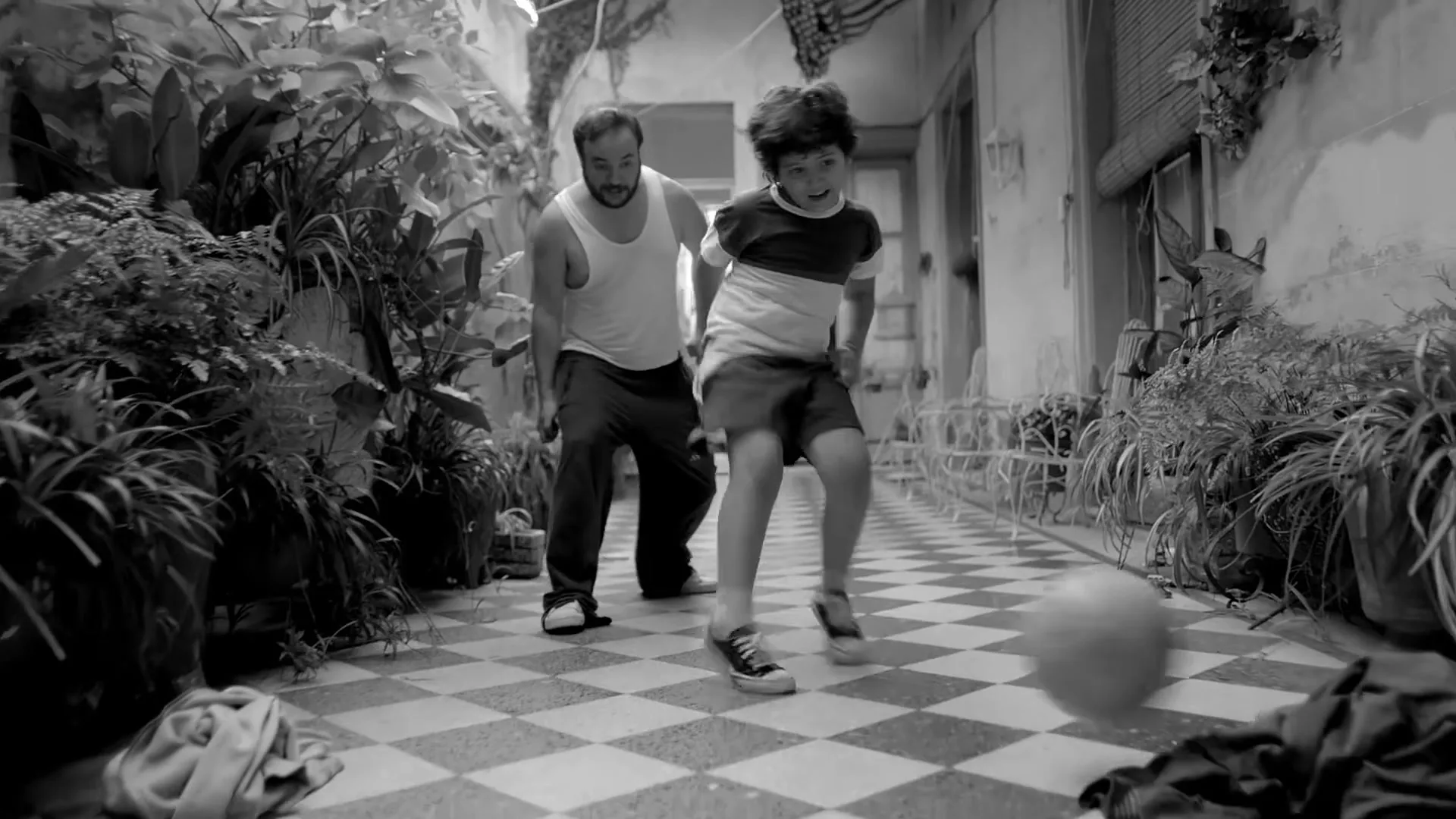 A father and son play soccer in a narrow, plant-filled hallway with a checkered floor, celebrating a goal with raised arms.