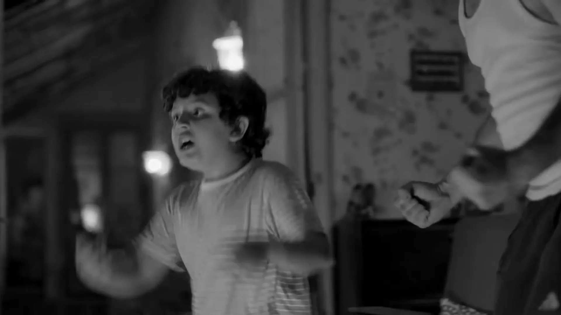 A young boy with curly hair, wearing a striped t-shirt, screams and gestures wildly while watching TV in a dimly lit living room.