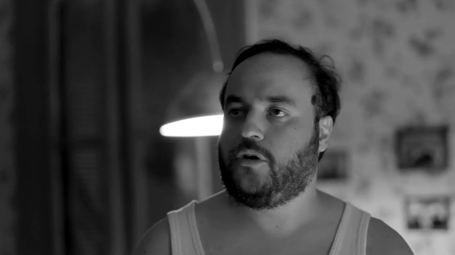 A man with a beard and receding hairline, wearing a tank top, looks surprised and slightly confused in a dimly lit room.