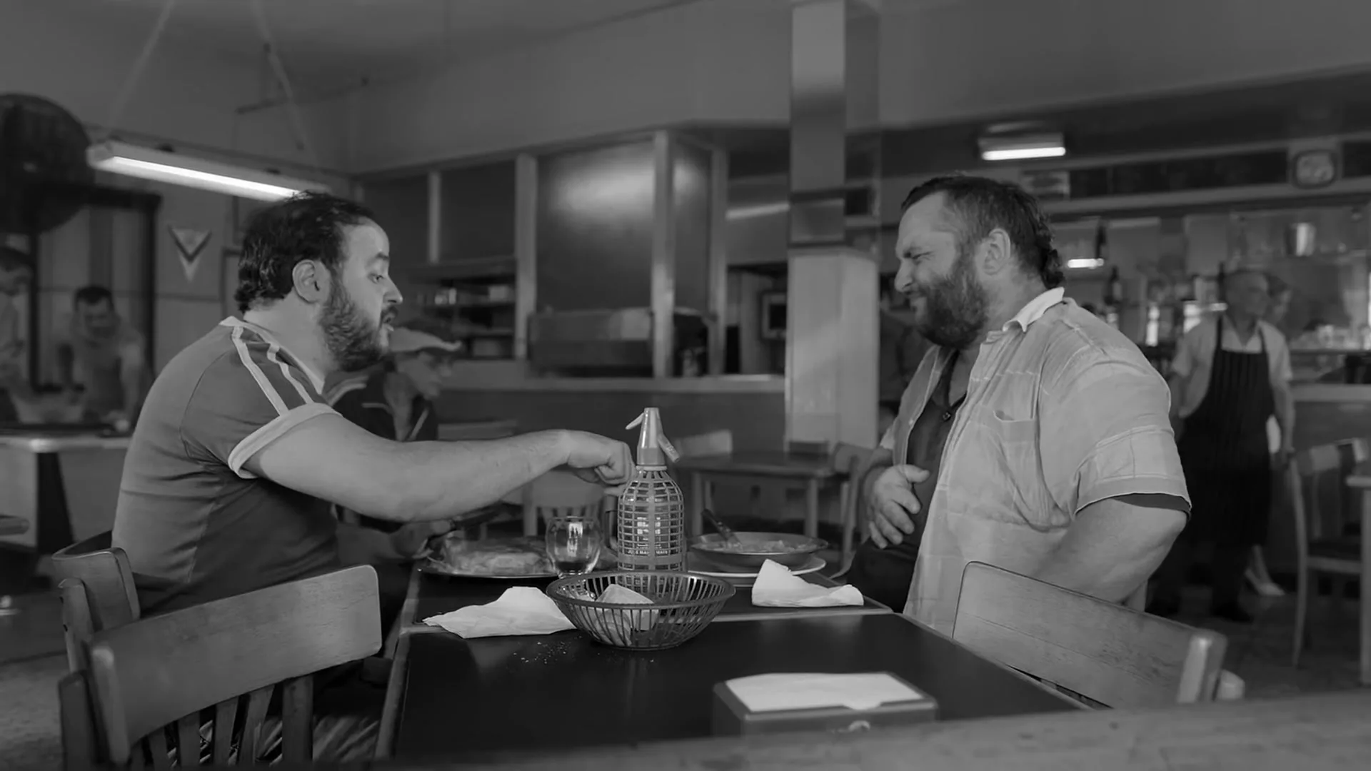 Two men with beards sit at a table in a diner, one points at a bottle while the other laughs, in black and white.