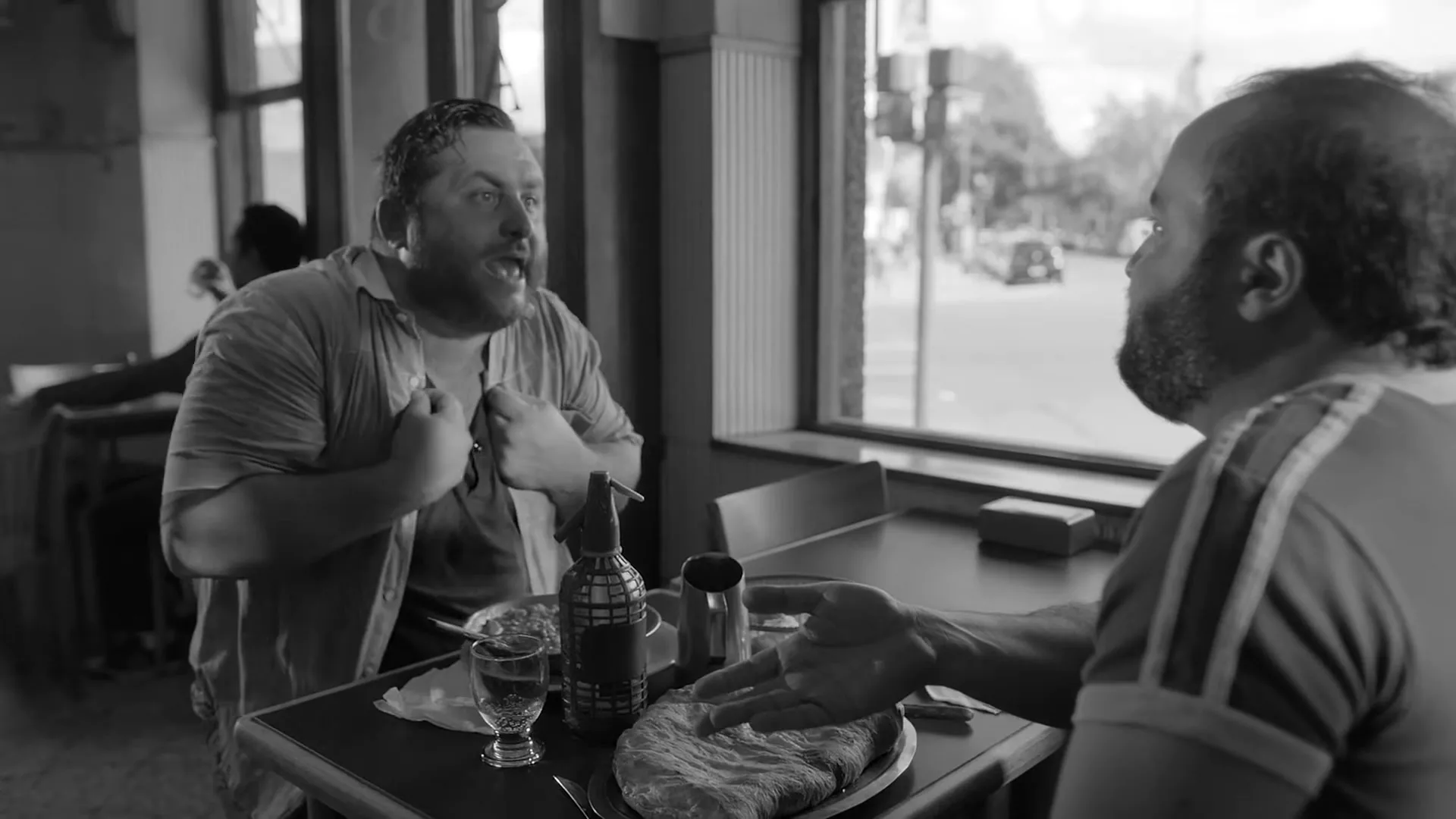 Two men with beards are sitting at a table in a diner, one gesturing emphatically while the other listens, in a black and white shot.