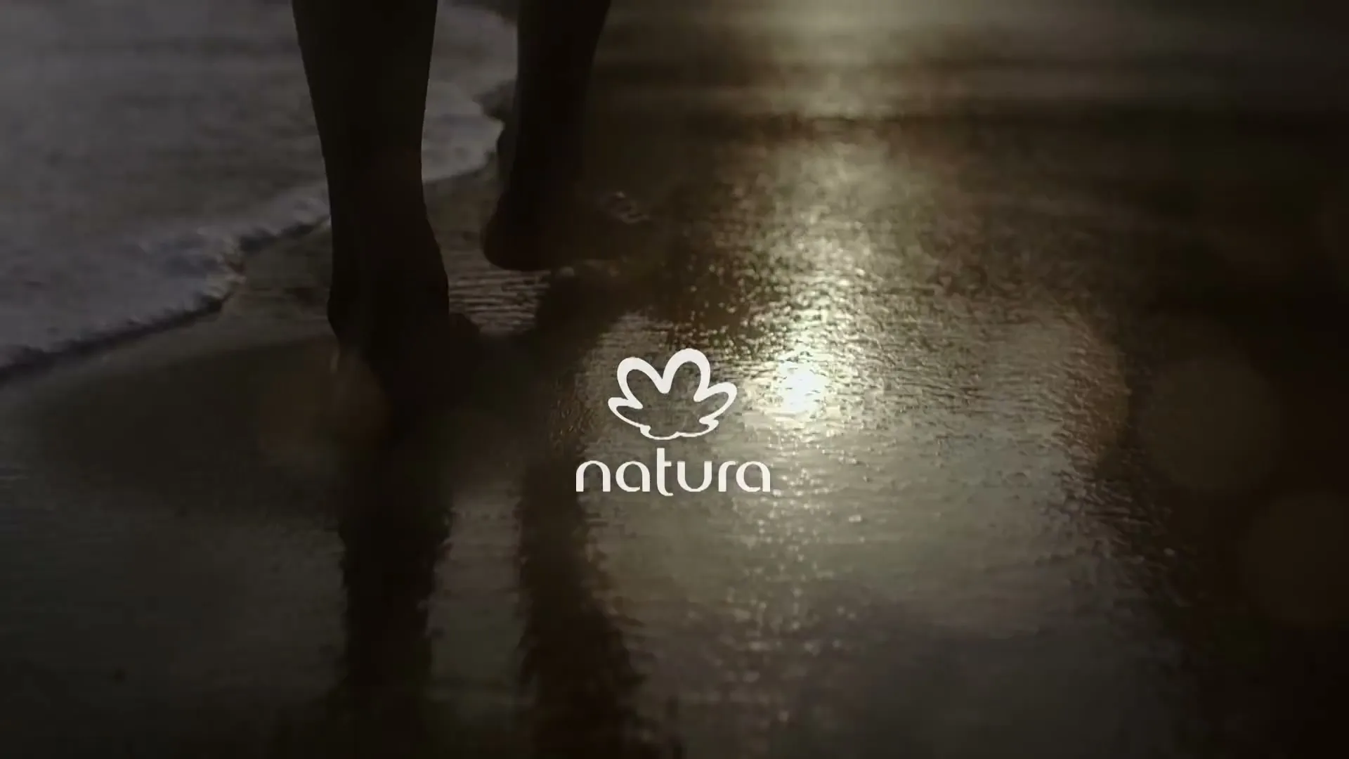 A person's bare feet walk on a wet beach at dusk, with the Natura logo superimposed over the scene.