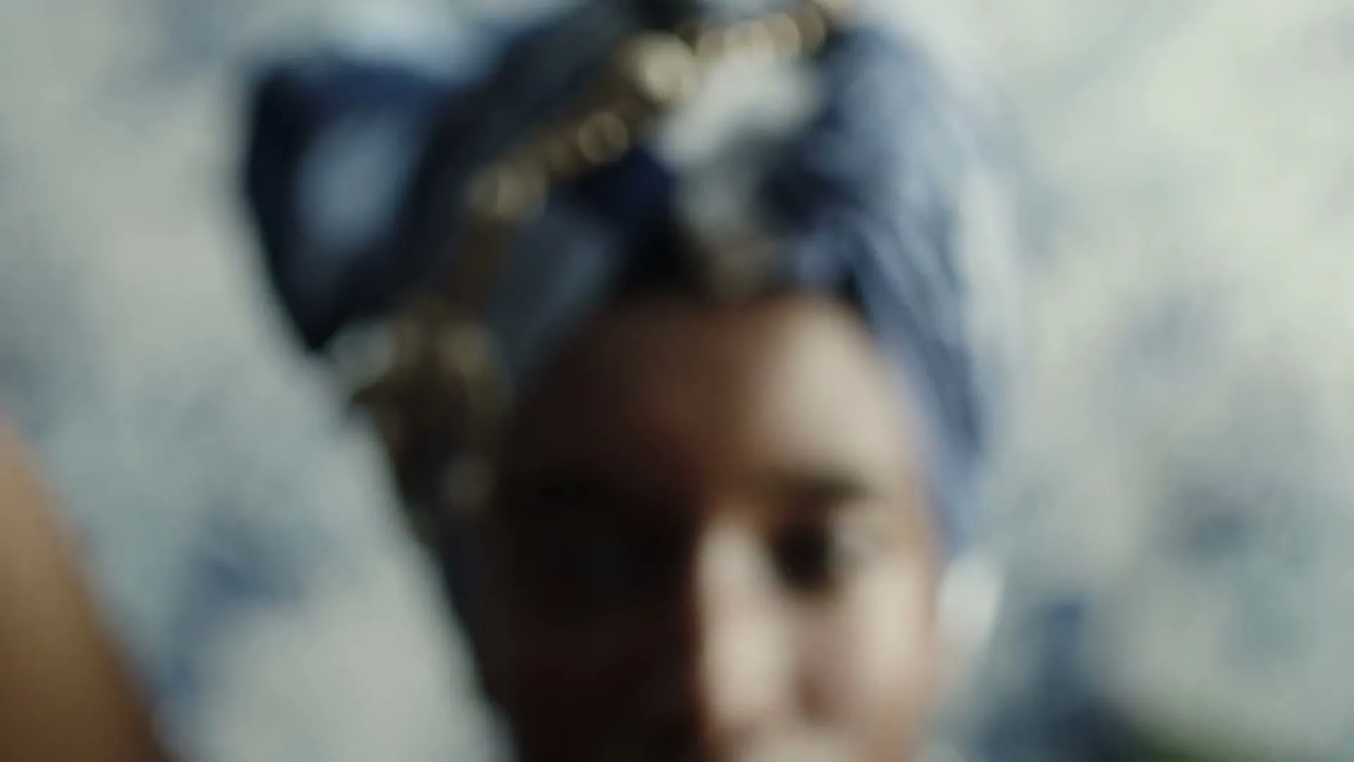 A woman with dark skin and a blue headwrap with gold jewelry looks directly at the camera as the shot comes into focus.