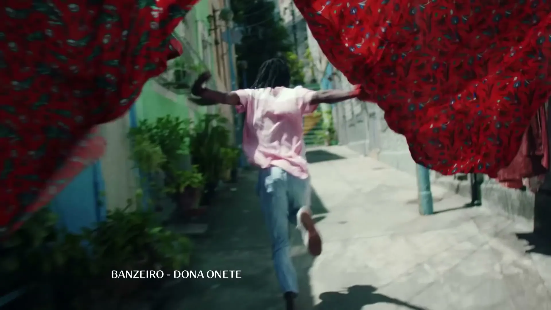 A young person with dreadlocks, wearing a pink shirt and jeans, runs down a narrow alleyway with colorful buildings and laundry hanging overhead.