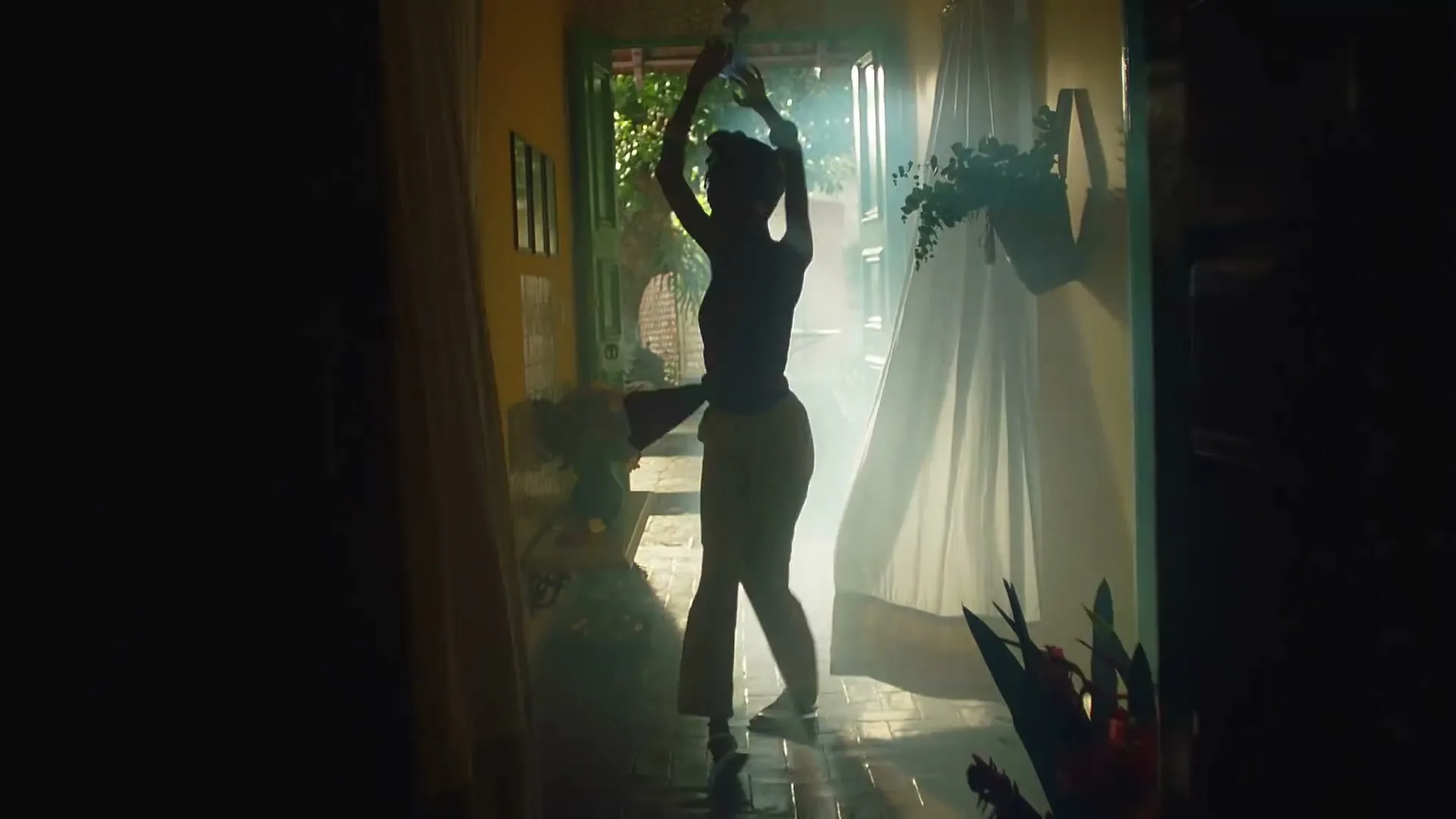 A silhouetted woman with arms raised dances in a doorway, with bright light and mist behind her, in a vibrant, plant-filled interior.