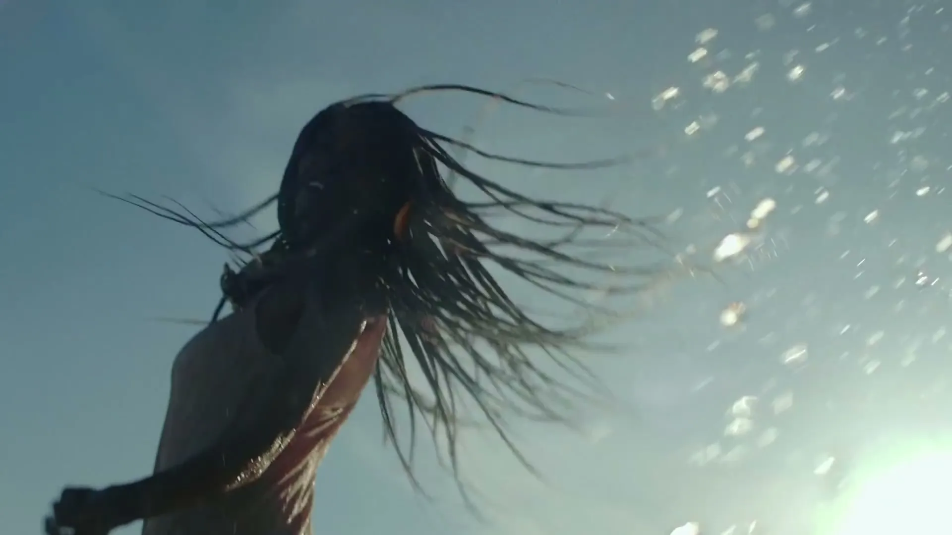 A young woman with long braids, wearing a tank top, is seen from a low angle, shaking her head as water splashes around her against a bright sky.