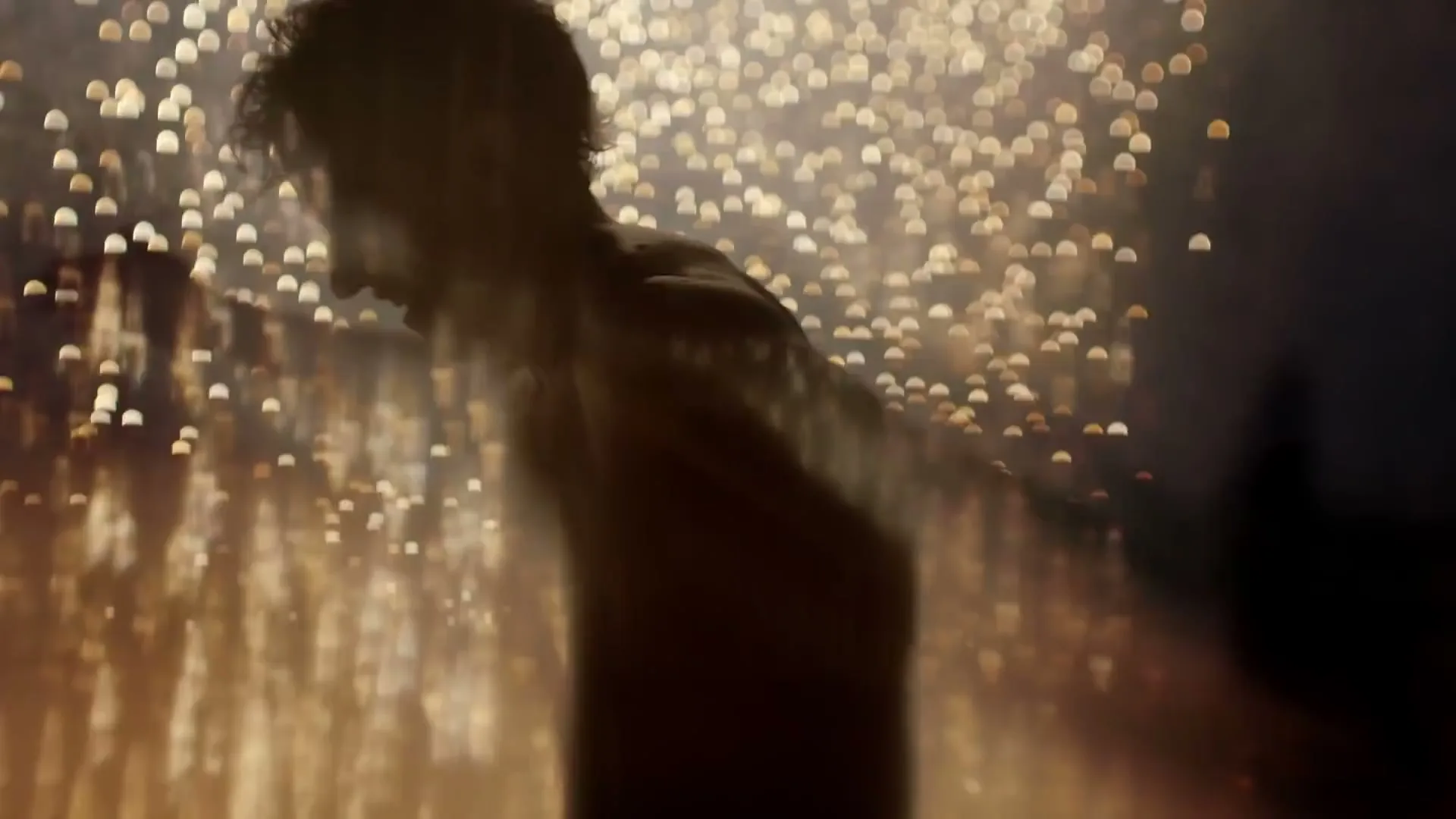 A shirtless man with curly hair is silhouetted against a background of sparkling lights, dancing at a party.