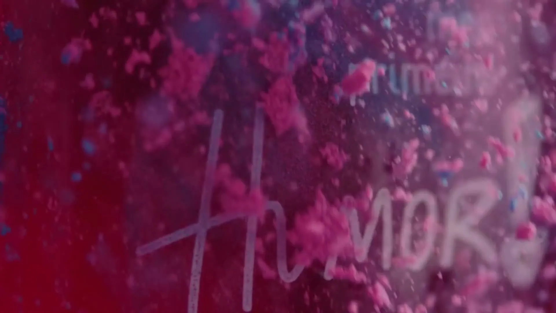 Pink and blue powder particles float and swirl around the word "Humor!" written in white on a dark pink background.