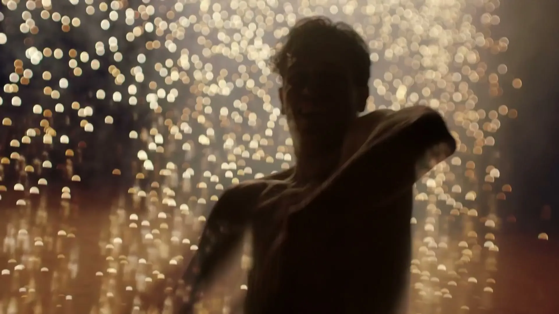 A shirtless man with curly hair dances in silhouette against a backdrop of sparkling golden lights, moving his arms gracefully.
