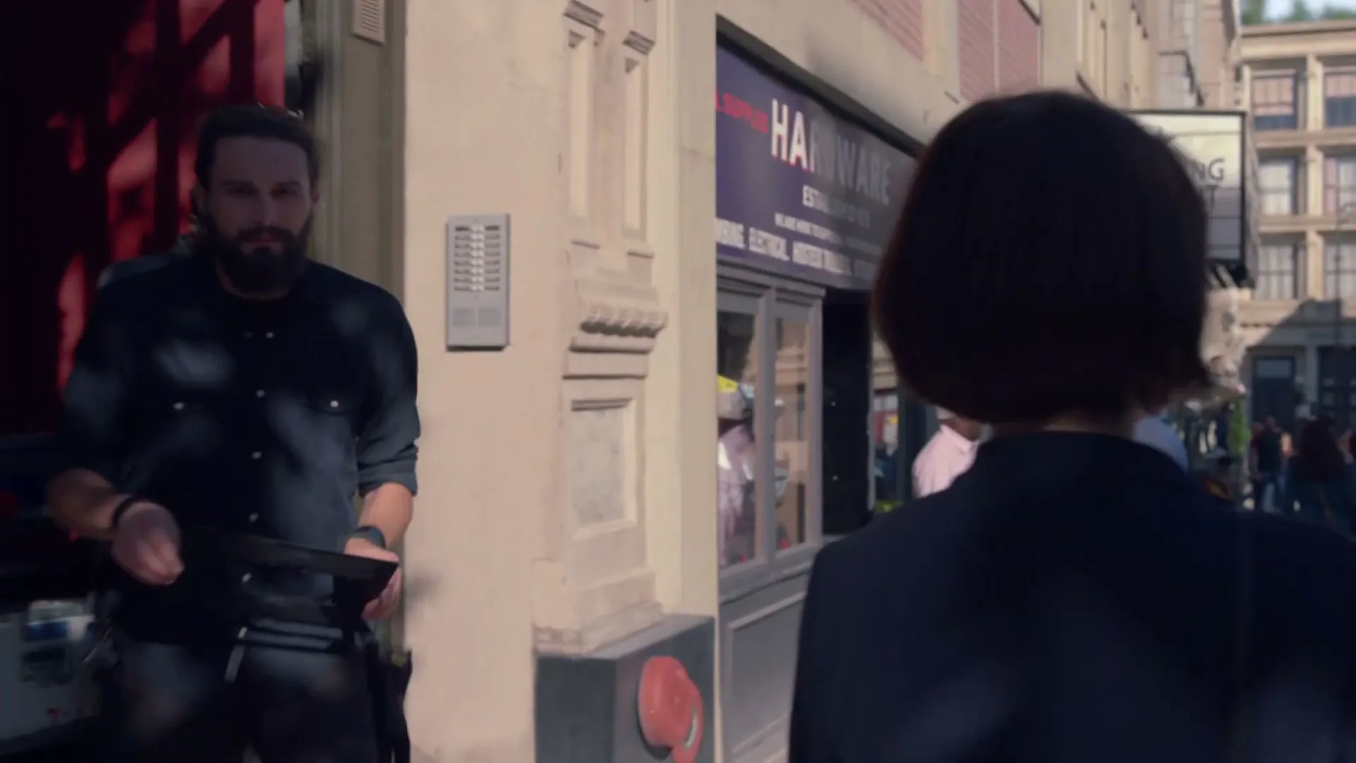 A woman with short dark hair walks down a city street, looking over her shoulder at a bearded man holding a camera frame.