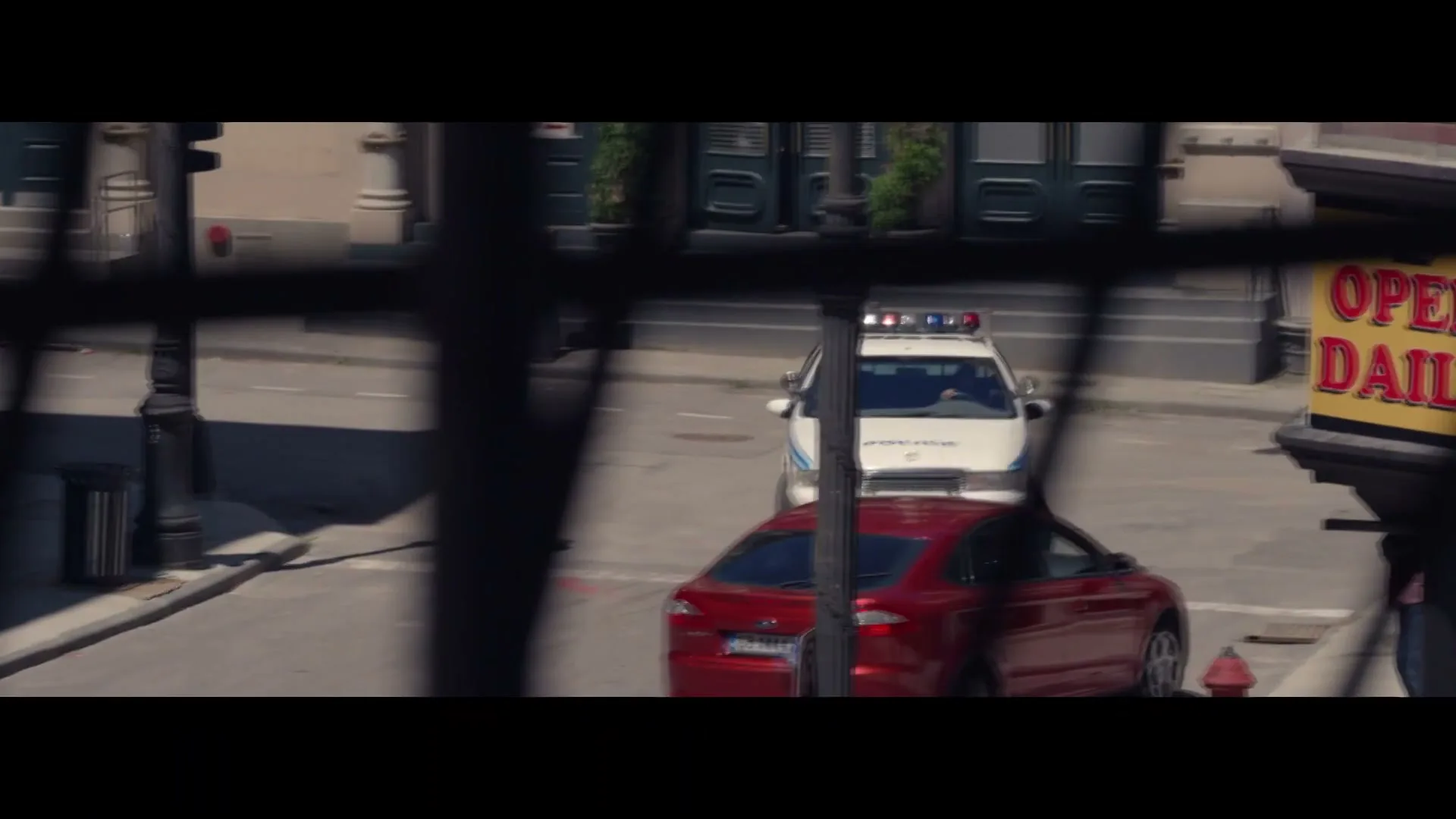 A red car and a police car drive down a city street, seen through a window with bars.